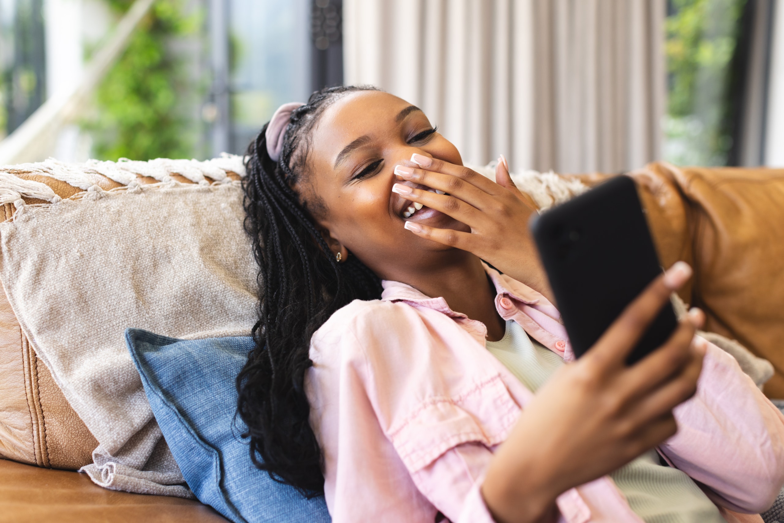 Young African American woman laughs while looking at her phone, reclining on a couch
